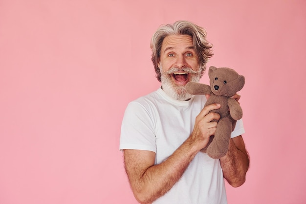 Hält Teddybär Stilvoller moderner Senior mit grauem Haar und Bart ist drinnen