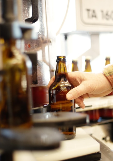 Hände Bier und Flasche in der Fabrikbrauerei oder Produktionsanlage zur Qualitätskontrolle Alkoholproduktionslinie und Person, die Glas von der automatischen Fördermaschine mit Produkt im Lager entnimmt
