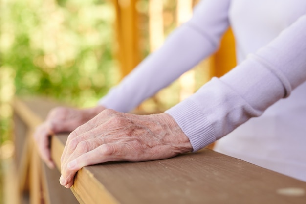 Hände der älteren Frau im weißen langärmeligen Pullover, der durch hölzerne Geländer ihres Landhauses während des Entspannens hält