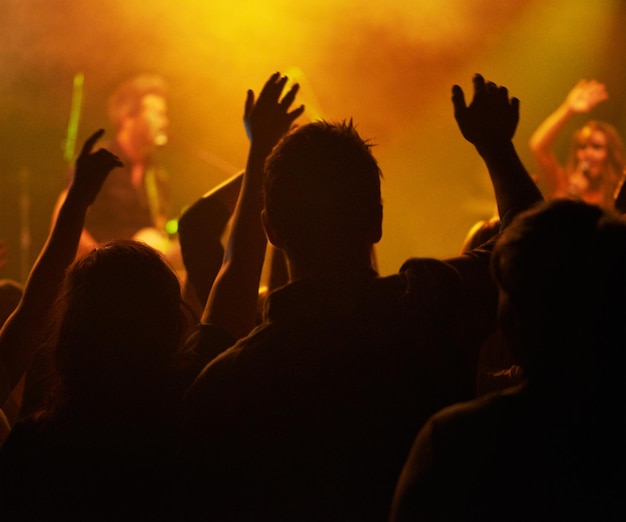 Hände der Fans und Silhouetten von Menschen bei Konzerten oder Musikfestivals von hinten, orangefarbene Lichter und Energie bei Live-Events. Tanzspaß und aufgeregtes Publikum in der Arena bei Rockband-Auftritten oder Publikumstanz
