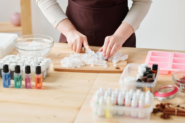 Hände der Handwerkerin, die transparente Seifenmasse an Bord durch Holztisch vor dem weiteren Behandlungsprozess schneidet