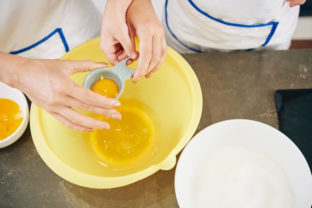 Hände der Mutter zeigen Tochter, wie man Plastikwerkzeug benutzt, wenn Eigelb von Eiweiß getrennt wird