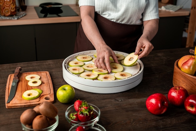 Hände der zeitgenössischen Hausfrau, die Scheiben frischer Äpfel auf eines der Tabletts mit Obsttrockner legen und gleichzeitig für den Winter sorgen