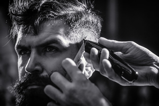 Hände des Friseurs mit Haarschneider. Barber arbeitet mit Haarschneidemaschine. Hipster-Kunde bekommt Haarschnitt. Haarschnitt-Konzept. Mann, der Friseur im Friseursalon besucht. Schwarz und weiß.