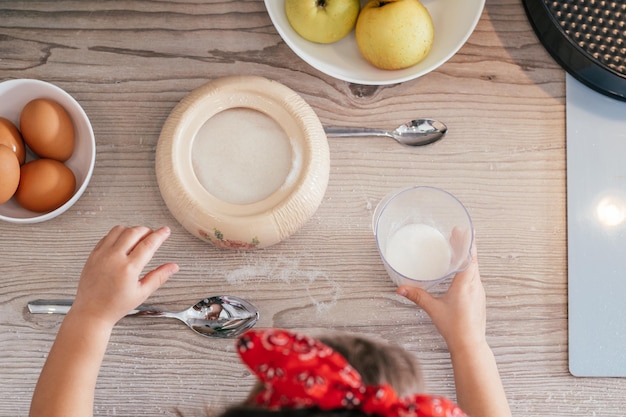 Hände des kleinen Mädchens im roten Stirnband backen Apfelkuchen in der Küche. Kind gibt Zucker in Messbecher. Kinder helfen bei der Hausarbeit. Kind, das Essen kocht. Ansicht von oben