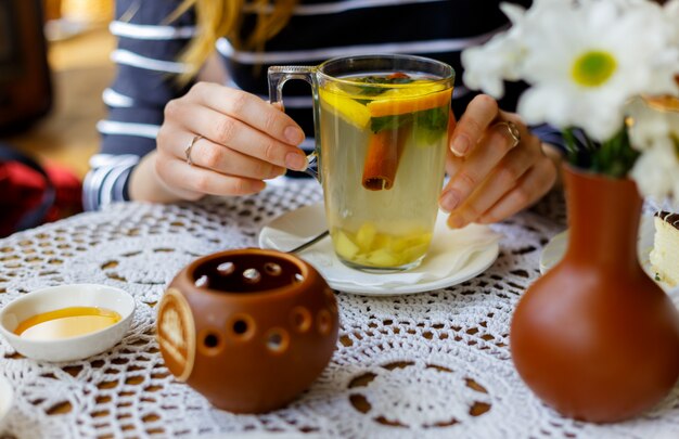 Hände des Mädchens halten eine transparente Tasse Tee mit Zimt, Minze, Orange auf dem Tisch mit Honig.