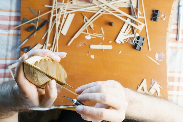 Hände des Mannes, der überschüssigen Kleber von Sperrholzdetails für Schiffsmodelle mit Gebrauchsmesser entfernt Prozess des Baus von Spielzeugschiffen