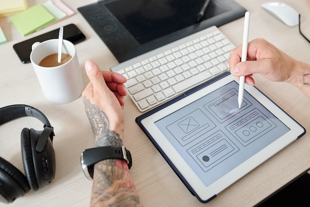 Hände des UI-Designers, der an der Schnittstelle einer neuen Website oder mobilen Anwendung arbeitet, zeigt er mit einem digitalen Stift auf einen Tablet-Computer