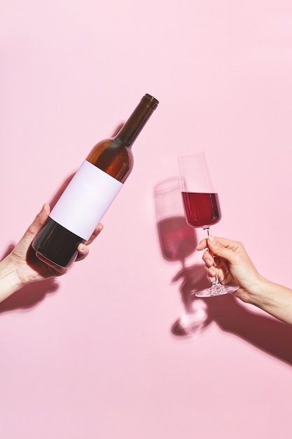 Foto hände, die ein glas wein und eine flasche auf rosa hintergrund halten