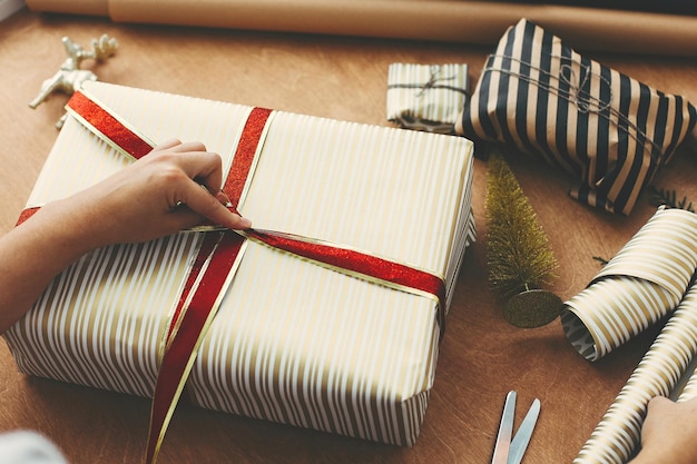 Foto hände, die ein rotes band halten und eine stilvolle geschenkbox in gestreiftes goldenes papier und eine schere einwickeln, präsentieren goldene baumhirsche auf einem holztisch konzept der weihnachtsgeschenke