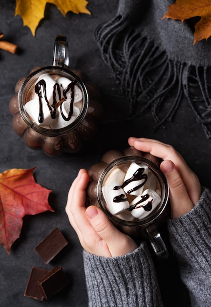 Hände, die eine Tasse heiße Schokolade mit Marshmallow auf einem dunkelgrauen Hintergrund mit Ahornblättern und Schal halten Herbstkonzept eines wärmenden Getränks Ansicht von oben xA