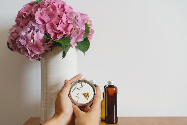 Hände, die festen Shampoo-Riegel oder natürliche Kräuterseife auf dem Hintergrund des Hortensienstraußes in der Vase und abfallfreie Hygieneartikel auf dem Holztisch im Zimmer halten Kunststofffrei