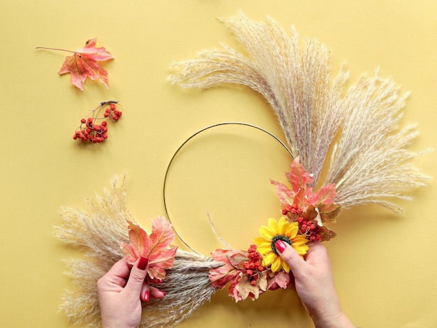 Hände, die getrockneten Blumenkranz aus trockenem Pampasgras und Herbstblättern machen, liegen flach auf gelbem Papier Sonnenlicht mit langen Schatten