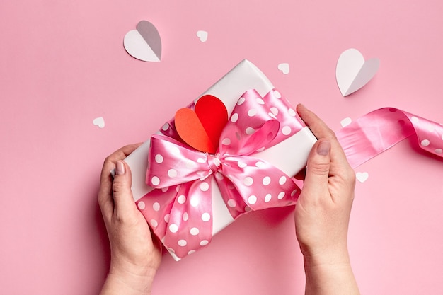 Hände, die Valentinstag-Geschenkbox mit Papierherzen auf rosa Hintergrund halten