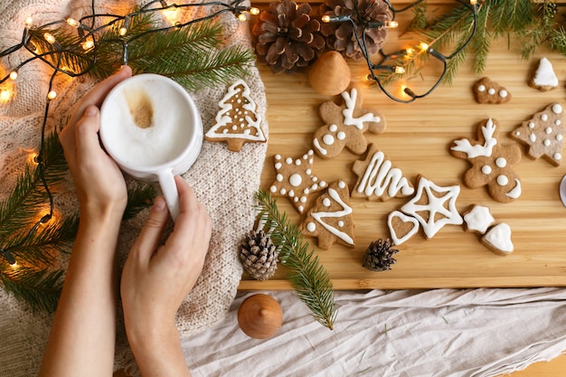 Hände, die warmen Kaffee auf dem Hintergrund von Weihnachtslebkuchen, gemütlichem Strickpullover, Tannenzweigen mit Tannenzapfen und Lichtern halten. Hallo Winter, stimmungsvolles Bild. Schöne Ferien!