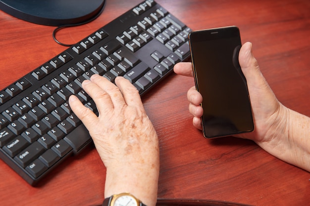 Hände einer älteren Frau, die auf einer Computertastatur tippt und ein Smartphone hält