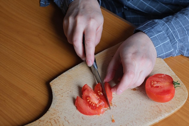 Foto hände einer älteren frau, die eine rote reife tomate auf einem schneidebrett für einen snack in der küche an einem braunen tisch schneidet, kein gesicht, nahaufnahme
