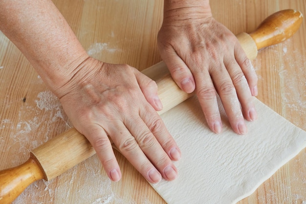 Hände einer älteren Frau rollen Teig zum Backen von Keksen mit einem Nudelholz aus.