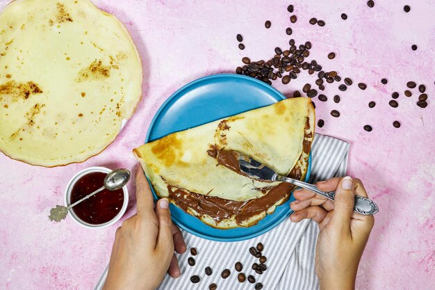 Hände einer Frau, die mit Schokoladenaufstrich gefüllte Crêpes auf einem blauen Teller mit Feigenmarmeladenschüssel und Kaffee isst