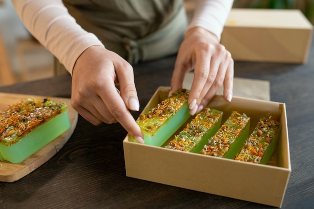 Hände einer jungen Frau, die frische handgemachte Naturseife in eine Karton-Geschenkbox verpackt, während sie vor dem Urlaub Geschenke für ihre Freunde vorbereitet