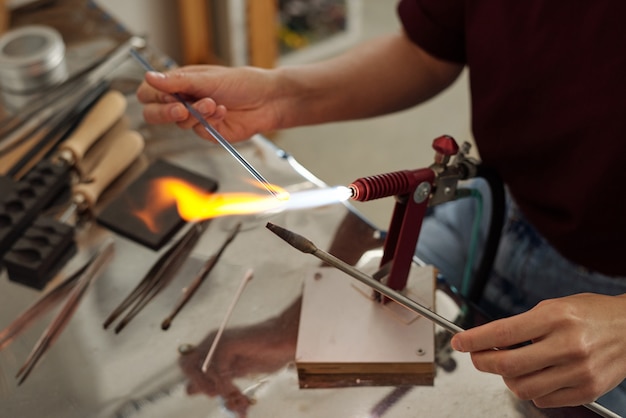 Hände einer jungen Lampenarbeiterin, die zwei Werkstücke über dem Brenner hält, während sie sie für die Weiterverarbeitung in der Werkstatt oder im Studio vorbereiten