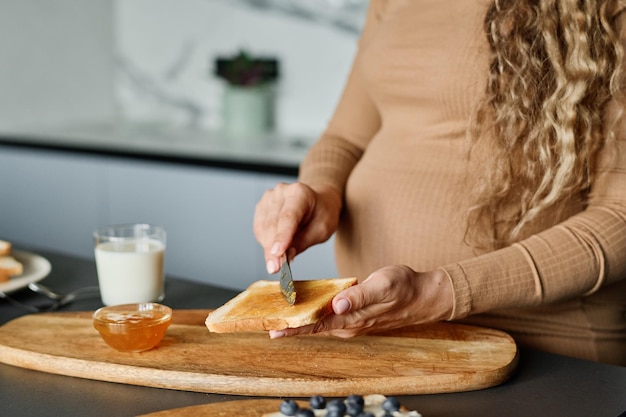 Hände einer jungen schwangeren Frau mit Messer, die Honig oder Marmelade auf Brot verteilt