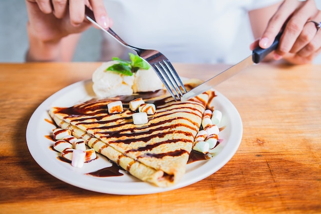 Hände einer Person mit einer Gabel, die Schokoladen-Crêpe und Eis schneidet. Frau isst Schokoladen-Crêpe und Eis mit einer Gabel