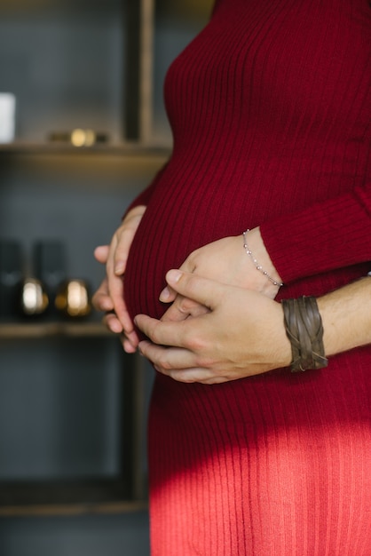 Hände einer schwangeren Frau und ihres Mannes auf seinem Magen in einem roten Kleid, Seitenansichtnahaufnahme