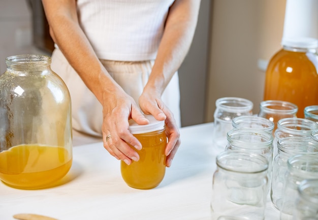 Hände einer unbekannten Frau schließen transparente Glasgefäße mit reinem süßem goldenem Honig mit Gummideckel, stehend auf einem großen weißen Holztisch und neben einer kleinen Untertasse mit einem Holzlöffel
