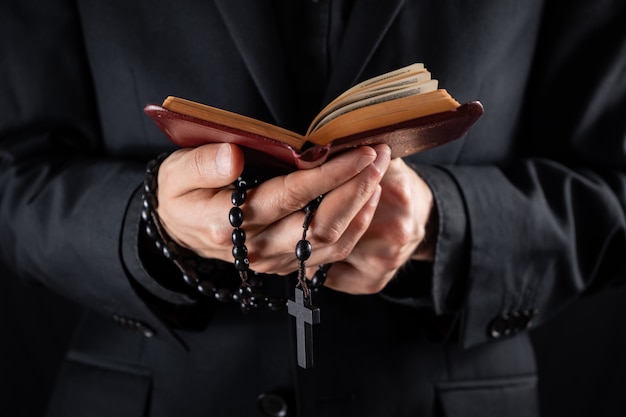 Hände eines christlichen Priesters kleideten im Schwarzen an, das ein Kruzifix hält und Buch des neuen Testaments liest. Religiöse Person studiert Bibel und hält Gebetsperlen, zurückhaltendes Bild