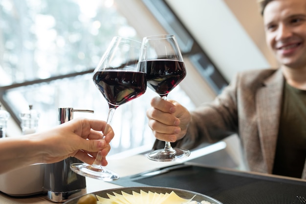 Hände eines glücklichen jungen Mannes und seiner Freundin, die beim romantischen Abendessen im Restaurant mit Gläsern Rotwein über dem servierten Tisch klirren