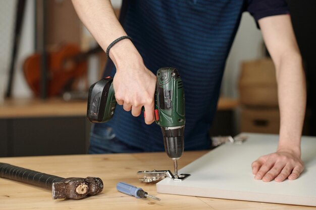 Hände eines Jungen, der eine elektrische Bohrmaschine hält, während er das Türscharnier des Küchenschranks repariert