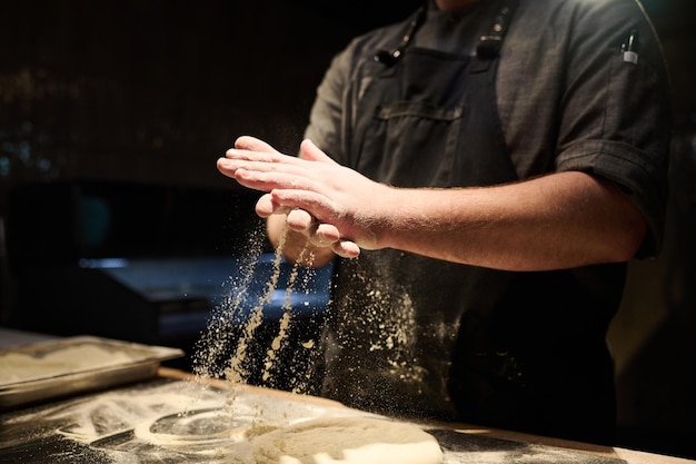 Hände eines jungen Kochs reiben Mehl zwischen den Handflächen und streuen es auf den Teig