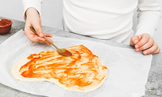 Hände eines kaukasischen Mädchens in einem weißen Rollkragenlöffel mit Tomatensauce auf einer herzförmigen Pizza