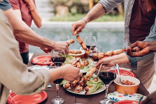 Hände greifen Spieße vom Tisch mit Weingläsern bei der Sommer-Barbecue-Party am Pool