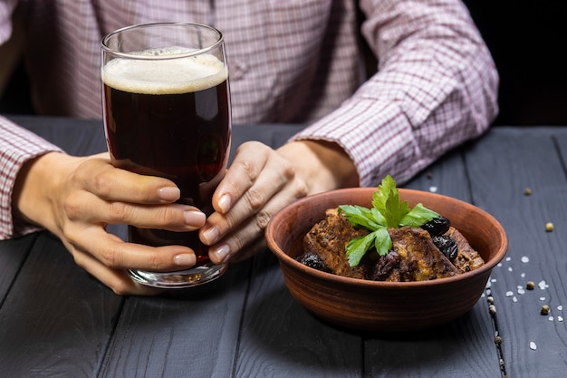 Hände halten Glas dunkles Bier und gegrillte Schweinerippchen mit frischem Petersilien-Zwetschgen auf schwarzem Holzhintergrund