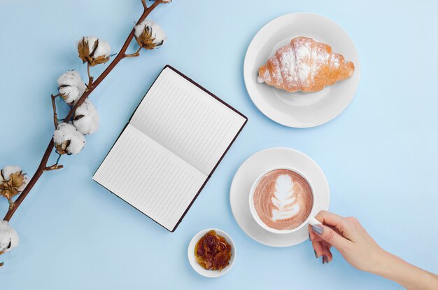 Hände halten Kaffee und Notizbuch mit Croissant und Baumwollzweig