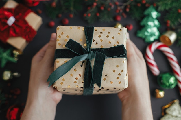 Hände halten stilvolles Weihnachtsgeschenk mit grünem Band auf schwarzem Hintergrund Frohe Weihnachten