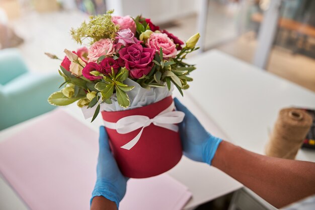 Hände in blauen Handschuhen halten einen Topf mit einem weißen Geschenkband mit rosa und roten Rosen rose