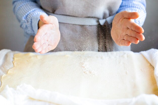 Hände in Mehl. Ein Hausbäcker in einer Schürze rollt den Teig und bestreut ihn mit Mehl