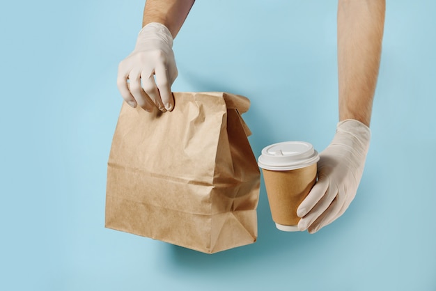 Foto hände in schutzhandschuhen, die eine papiertüte und eine kaffeetasse halten