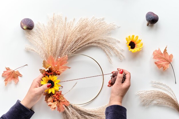 Hände machen getrockneten Blumenkranz aus trockenem Pampasgras und Herbstlaub Hände binden Dekorationen an Metallrahmen Flach auf weißem Tisch weiches natürliches Sonnenlicht