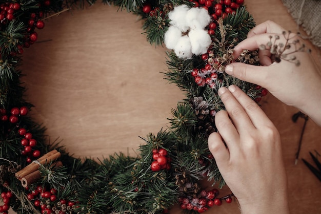 Foto hände machen rustikalen weihnachtskranz mit kräutern an tannenzweigen rote beeren tannenzapfen baumwolle auf rustikalem holzhintergrund atmosphärisches stimmungsvolles bild im winterurlaubsworkshop