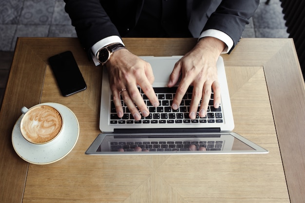 Hände Männer liegen auf dem Laptop, in der Nähe eines Telefons und Cappuccino