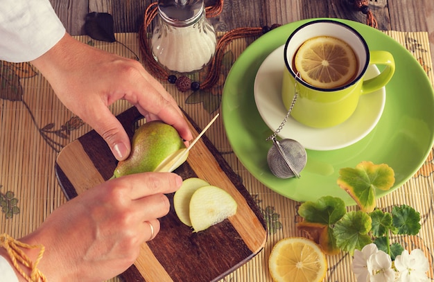 Hände schneiden eine Birne. Nächster Tee mit Zitrone in einer grünen Tasse, Zuckerdose, einem Brett mit einer grünen Birne und einem Messer. Stimmungsvolles Foto. Frische helle Farben. Ansicht von oben.