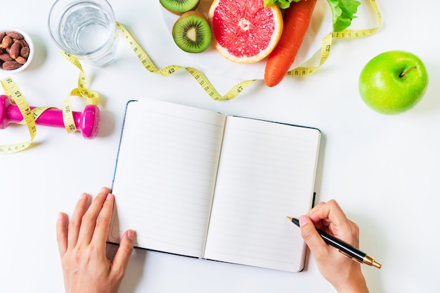 Hände schreiben auf Notizbuch mit Obst, Gemüse, Hantel, Maßband und Wasser. Sauberes Essen und Bewegung für ein gutes Gesundheitskonzept. Bio-Lebensmittel, Diätkonzept. Draufsicht,