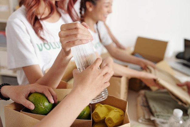 Hände von Freiwilligen des Wohltätigkeitszentrums, die Lebensmittel, Wasser und frisches Obst in Kartons packen