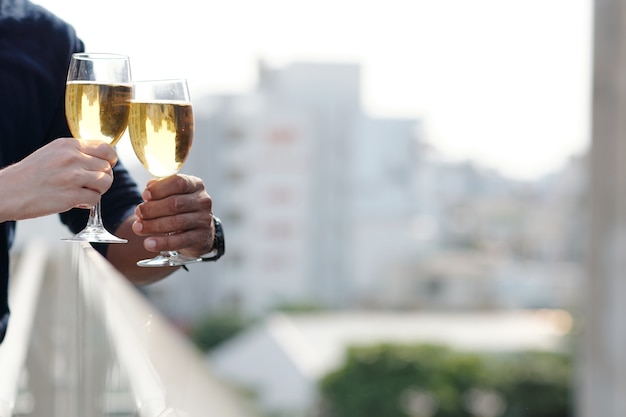 Hände von Mann und Frau, die beim Stehen auf dem Balkon mit kaltem Weißwein anstoßen, Großstadt im Hintergrund