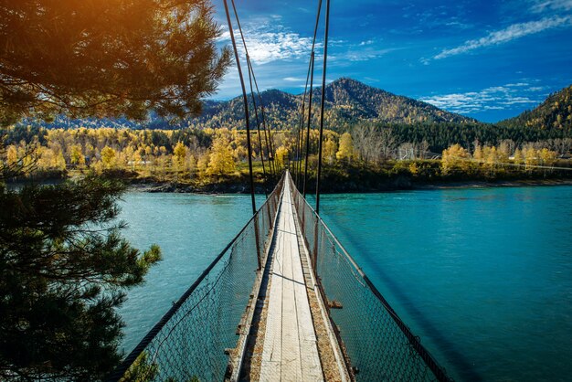 Hängende Holzbrücke des Fußgängers über Gebirgsfluss. Lange Brücke über den Fluss gegen Berge bedeckt mit Wald, goldenem Herbst, warmem sonnigem Tag des indischen Sommers