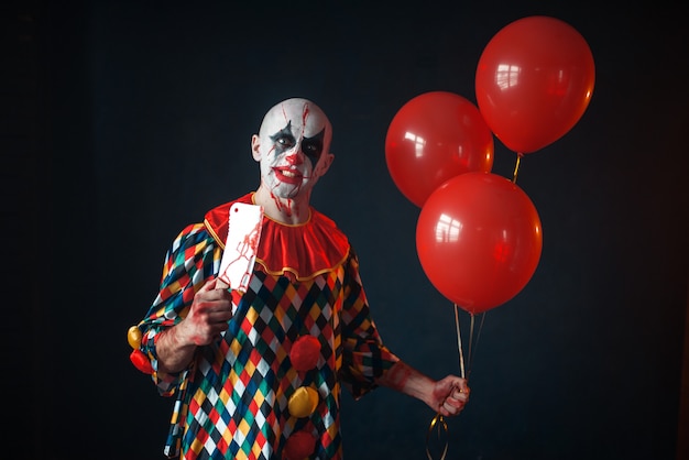 Hässlicher blutiger Clown mit Fleischerbeil hält Luftballon, Entsetzen. Mann mit Make-up im Karnevalskostüm, verrückter Verrückter